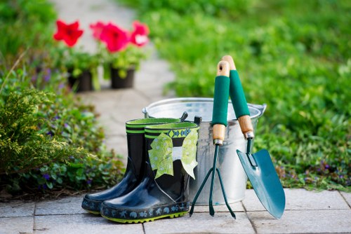 An outdoor garden scene featuring a paved area with garden tools and accessories arranged on the ground. Prominently, a pair of black and green rubber gardening boots with a leafy pattern are positioned next to a white plastic bucket. Inside the bucket, there are gardening tools including a hand trowel with a blue handle and a long-handled weeder. To the right of the bucket, a metal gardening fork with a green handle rests against the bucket, and a small blue trowel is embedded into the ground nearby. The background reveals a lush, green garden with vibrant pink petunias in pots, along with various shrubs and a grassy lawn. The overall scene suggests preparation for gardening or lawn maintenance, with natural daylight illuminating the setting, indicative of a clear, pleasant day in a residential outdoor space, typical of gardens serviced by Lawn Mowing Temple in the local area of Temple or nearby regions.